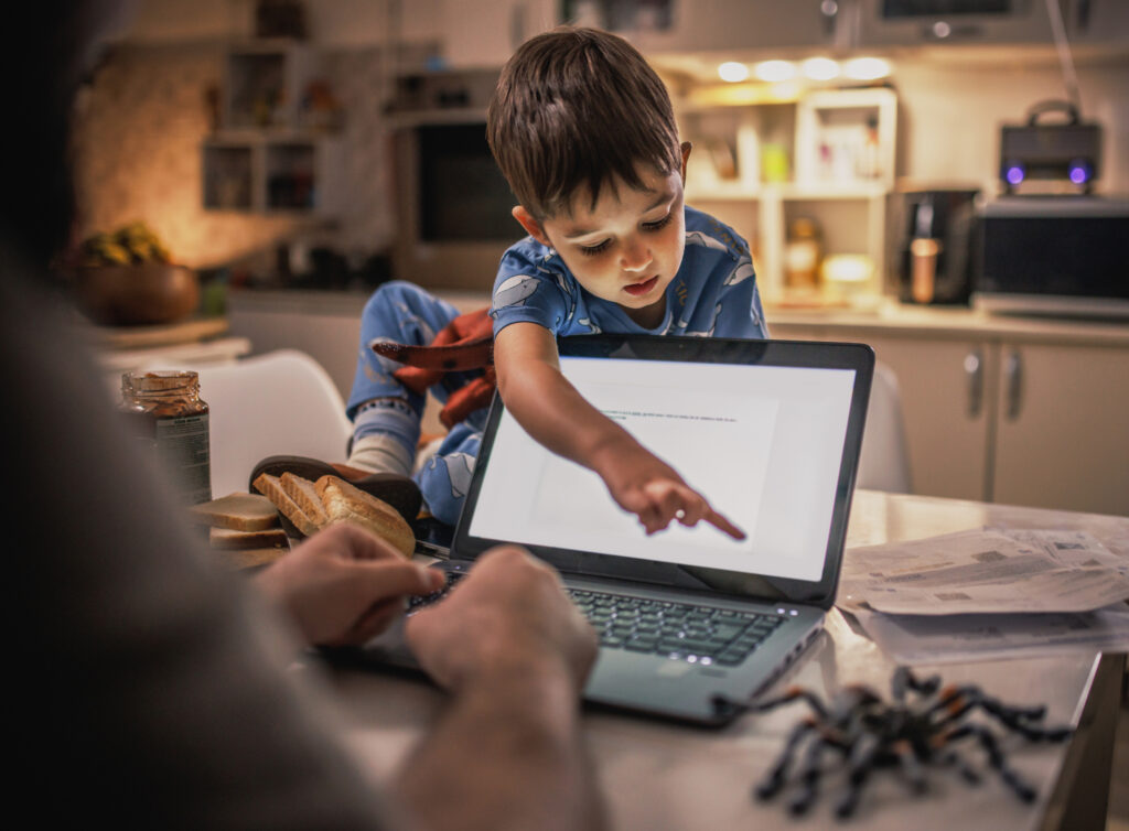 Young boy in pajamas sits on a kitchen counter pointing at a laptop screen with interest while an adult types nearby, creating a cozy scene that relates to foster care and taxes in a warm, supportive home environment.