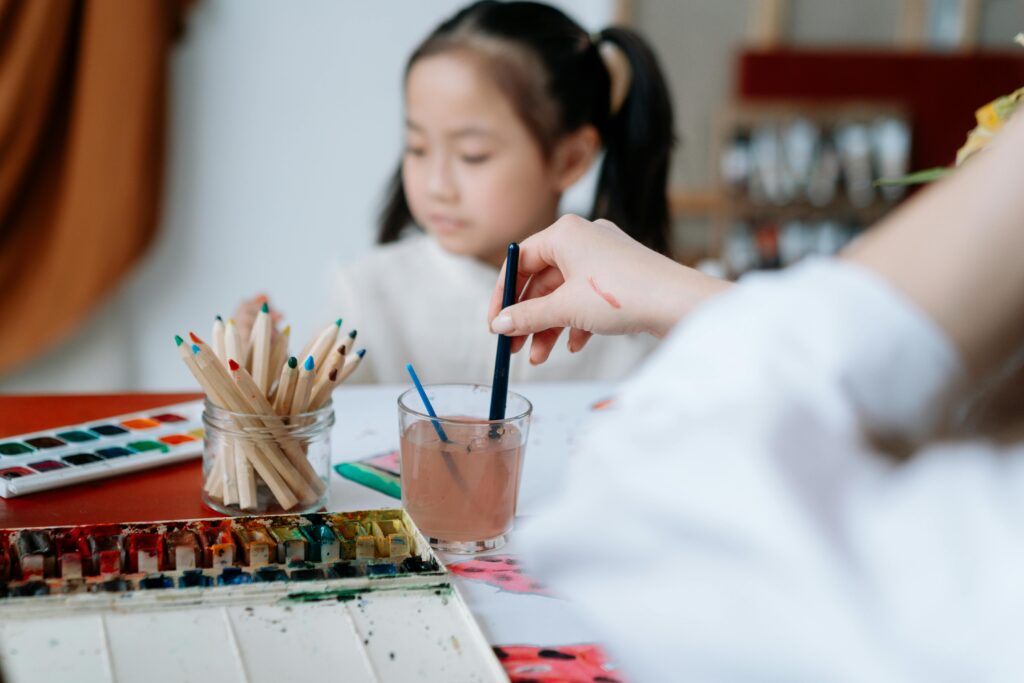 A child focuses on drawing at a table filled with art supplies, illustrating the different types of foster care. Nearby, a hand rinses a paintbrush in a jar of water, creating a creative and peaceful atmosphere.