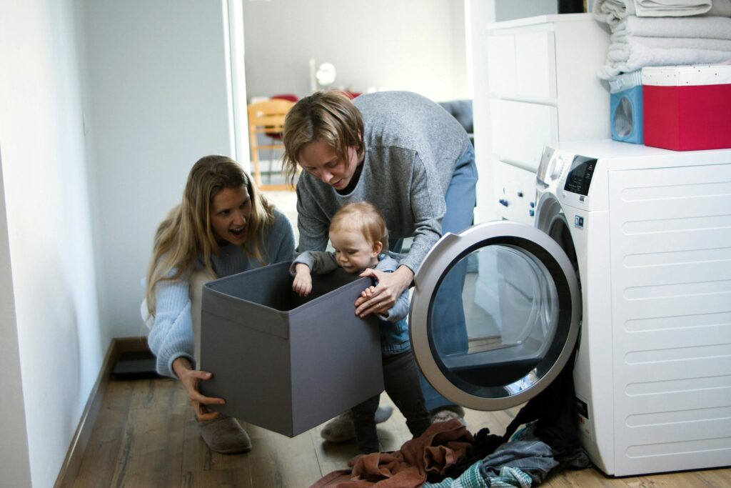 A joyful scene of a family in a laundry room experiencing temporary foster care. A young child climbs into a box held by two adults next to a washing machine. Towels are stacked nearby.