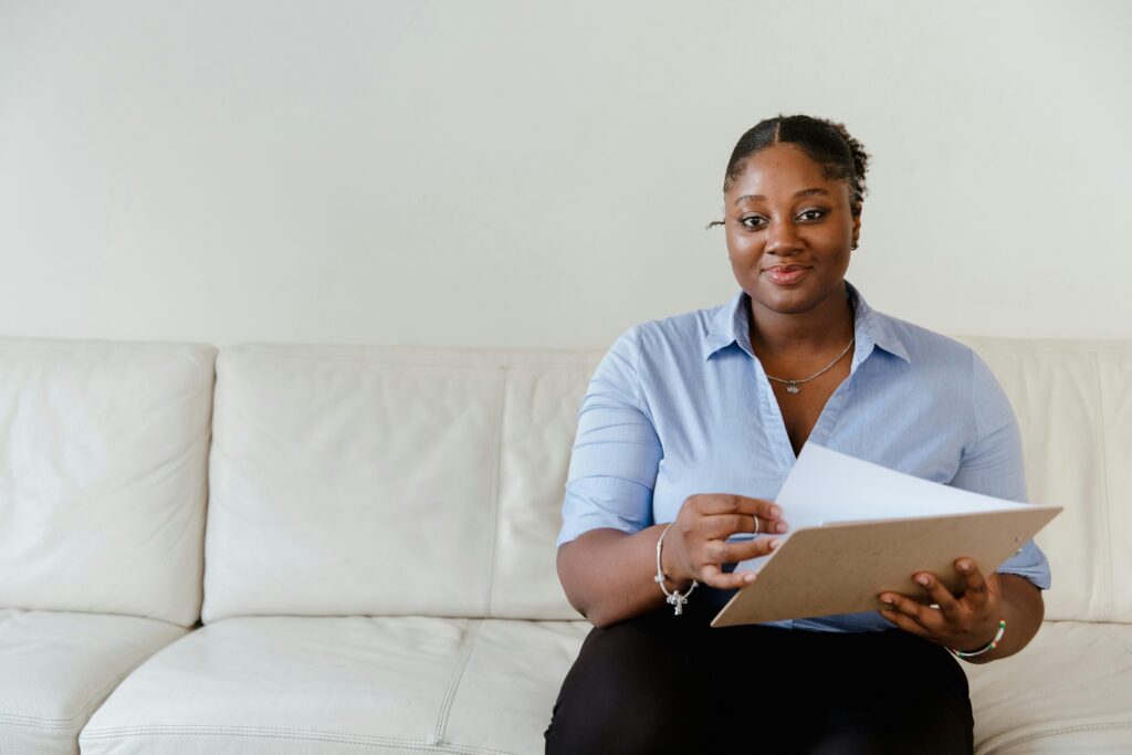 A person sits on a white sofa, holding a clipboard and papers, smiling softly in a welcoming and professional manner, representing free foster parent training.