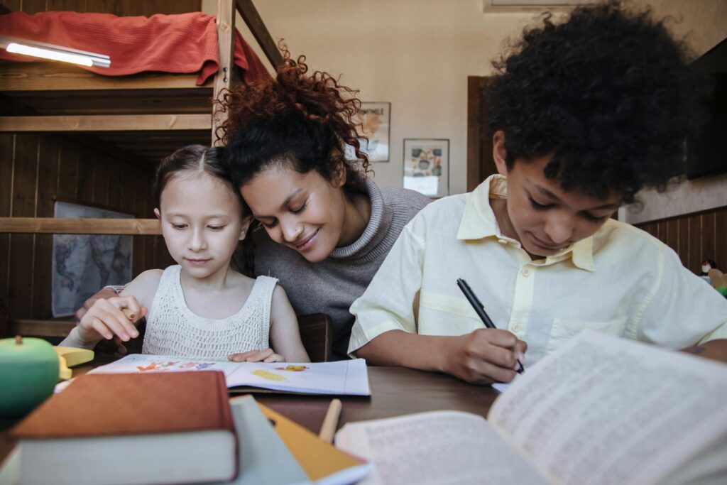 A woman and two children sit at a table, focused and smiling, engaged in learning activities with Every Child TN. One child colors while the other writes. The atmosphere is warm and supportive.