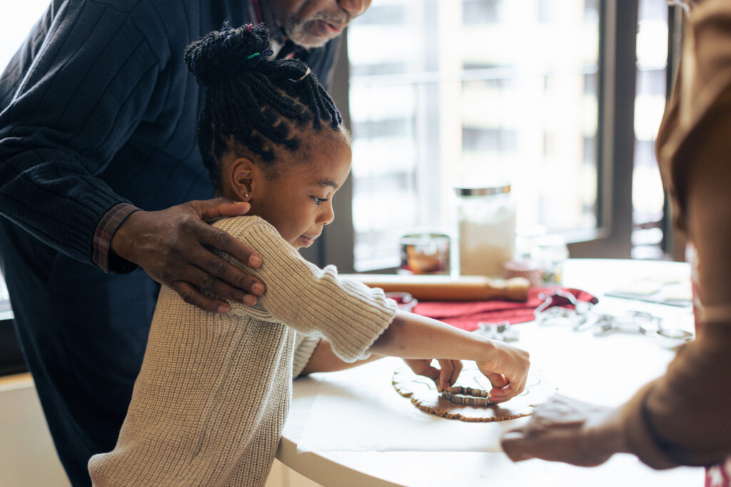 A young girl with braided hair uses a cookie cutter on dough, guided by an older man who gently places a hand on her shoulder. The sunlit kitchen setting conveys warmth and highlights the joy of fostering during the holidays.