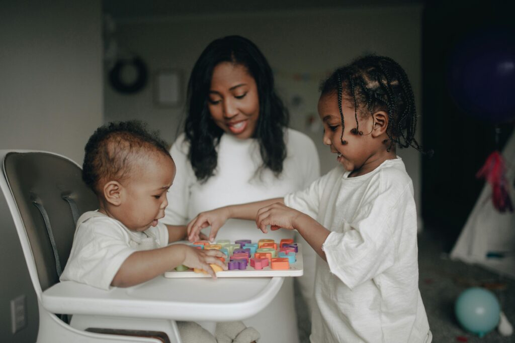 A caregiver smiles while two young children play with a colorful puzzle on a white highchair tray, illustrating a warm and supportive Respite Care environment.