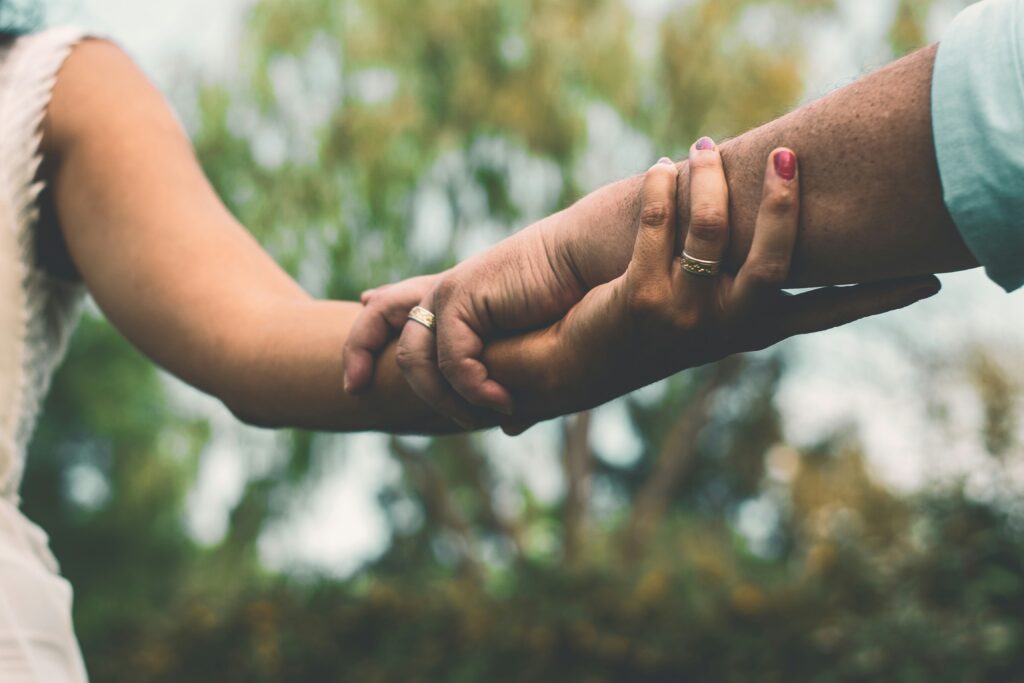 Two people holding hands outdoors, one with pink nail polish, symbolizing cultural continuity and connection against a backdrop of green trees.