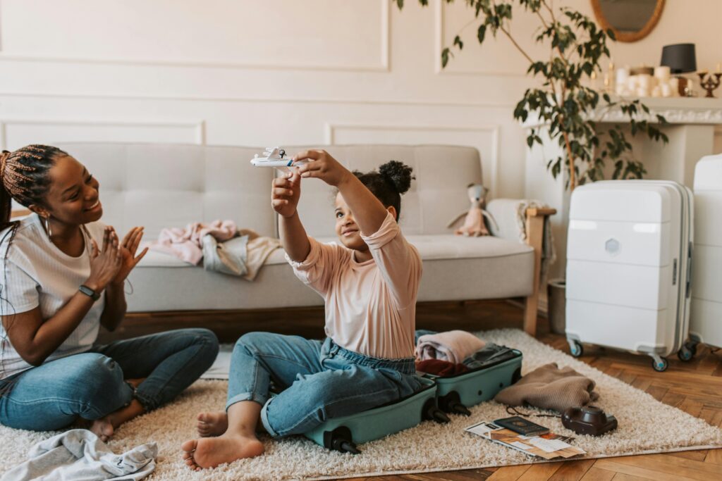 A child sits inside an open suitcase playing with a toy airplane, while an adult, sitting on the floor, watches and claps. The room has a cozy, warm ambiance.