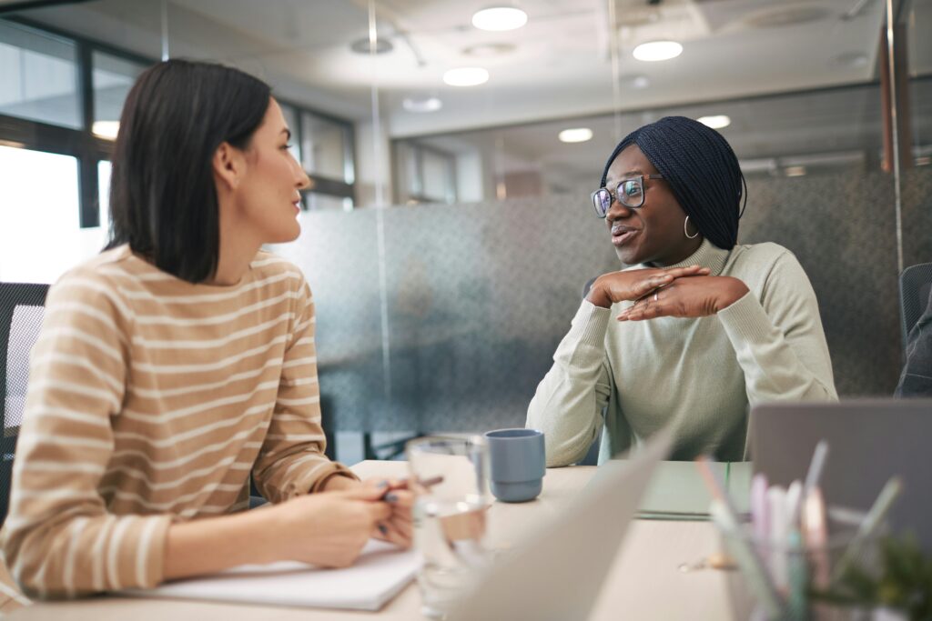 Two women sit at a conference table with laptops and mugs, engaged in conversation, highlighting the importance of case workers in supporting families.