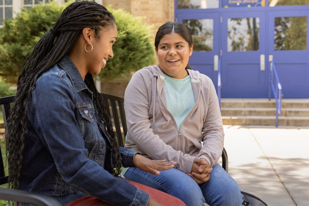 Two women, one in a denim jacket and one in a hoodie, chat on an outdoor bench, illustrating the positive impact of CASA volunteers in the community.