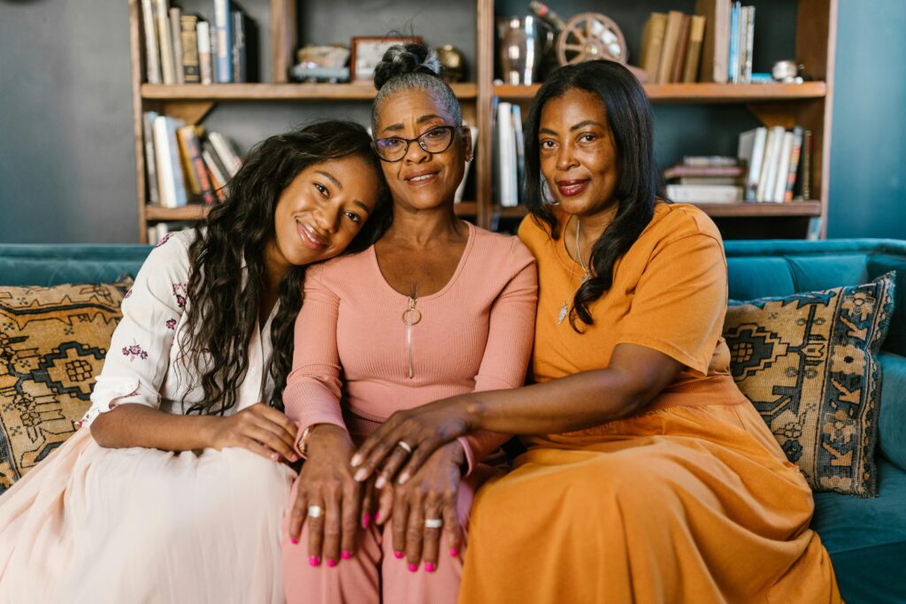 Three women sit closely on a couch, conveying warmth and togetherness, highlighting the importance of family reunification. A young woman leans on the older woman's shoulder. Bookshelves in the background.