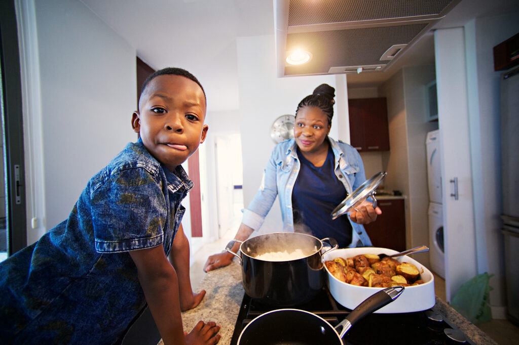 Foster mother and son busy at in the kitchen with the boy looking at the camera licking his lips while the mother looks and smiles.