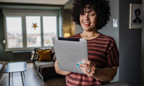 Woman with curly hair smiling at a tablet in a cozy living room. Natural light from large windows, star decoration visible outside, serene atmosphere.
