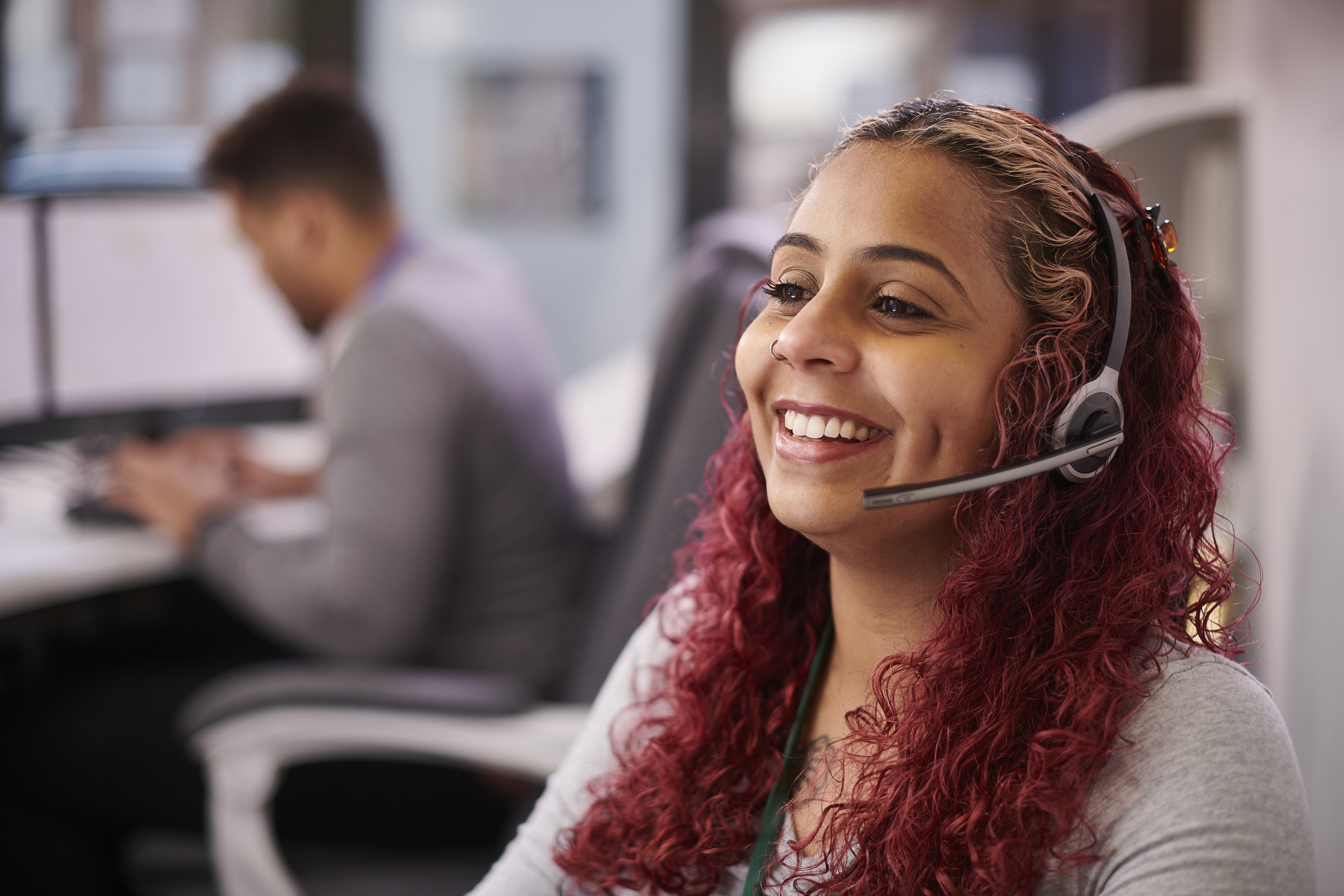 Smiling woman with curly red hair wearing a headset sits at a desk, suggesting a call center environment. The background shows a blurred colleague working. 