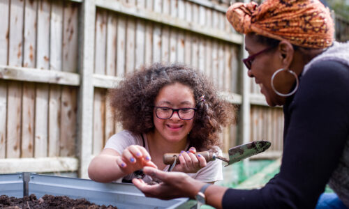 A young girl with glasses digs in a garden bed, smiling at a woman handing her a seed. They are enjoying the day in a fenced backyard.