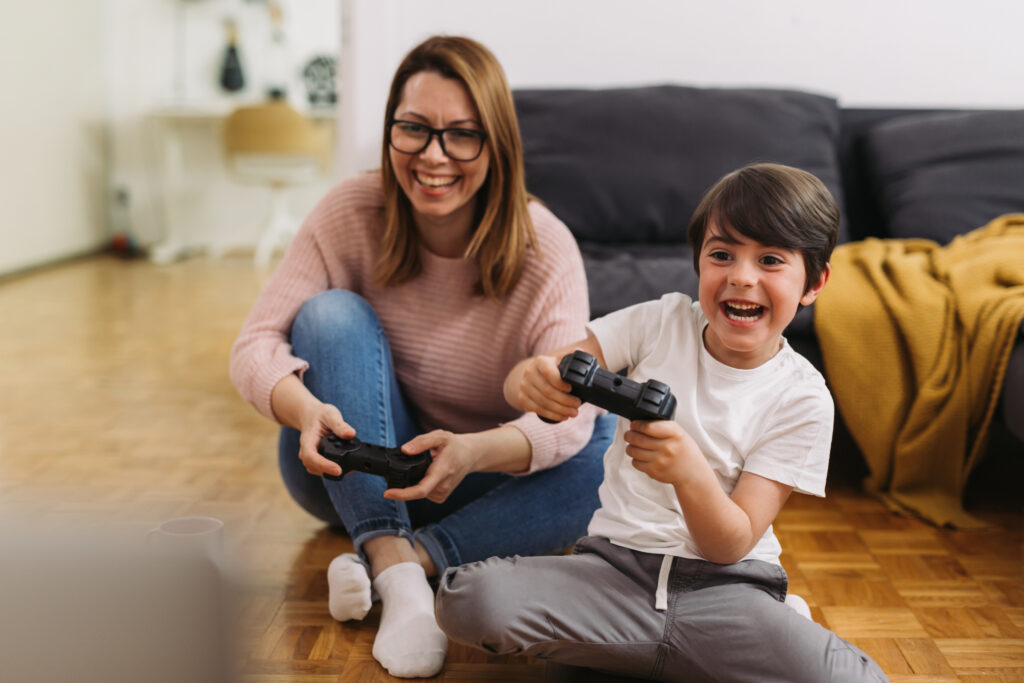 A foster mother and son happily play video games together on the floor. They are sitting near a sofa, holding controllers, experiencing joy and excitement.